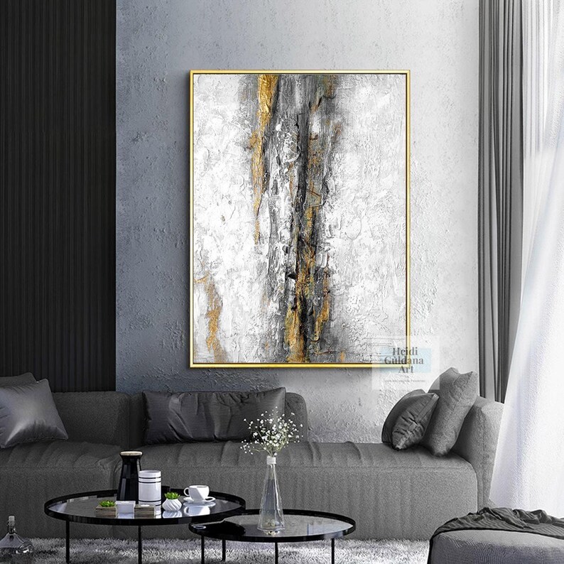 May include: Abstract painting with gold, gray, and white colors. The painting is framed in a gold frame and is hanging on a gray wall.