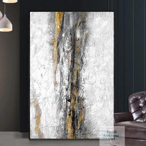 May include: Abstract painting with gold, black, and white paint on a white canvas. The painting has a textured look and is framed in a black frame.