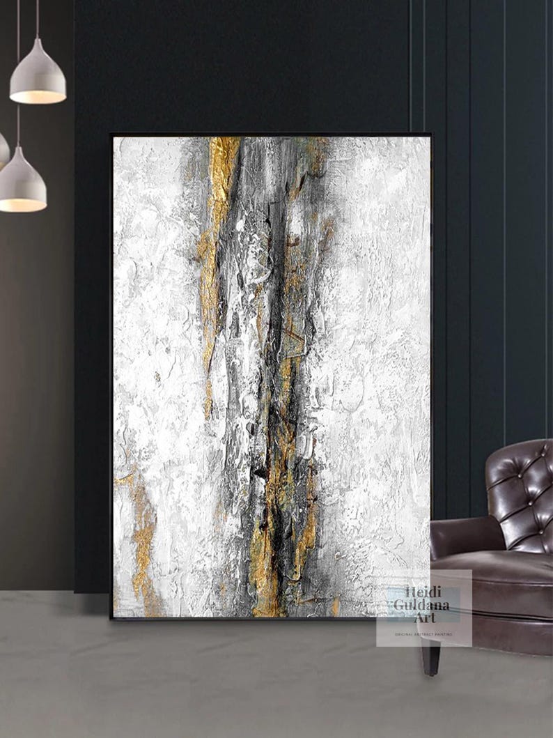 May include: Abstract painting with gold, black, and white paint on a white canvas. The painting has a textured look and is framed in a black frame.