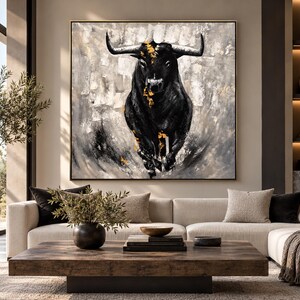 May include: A square wall art piece featuring a charging black bull centered against a textured gray and white background. The bull is accented with small, vibrant gold leaf details along its head and chest, framed in a thin, dark metal border.
