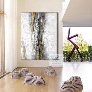 May include: Abstract painting in shades of white, gray, and gold with black accents. The painting is hanging on a white wall in a modern interior with a purple metal sculpture in the background. There are four woven, purple and white, round, decorative objects on the floor.