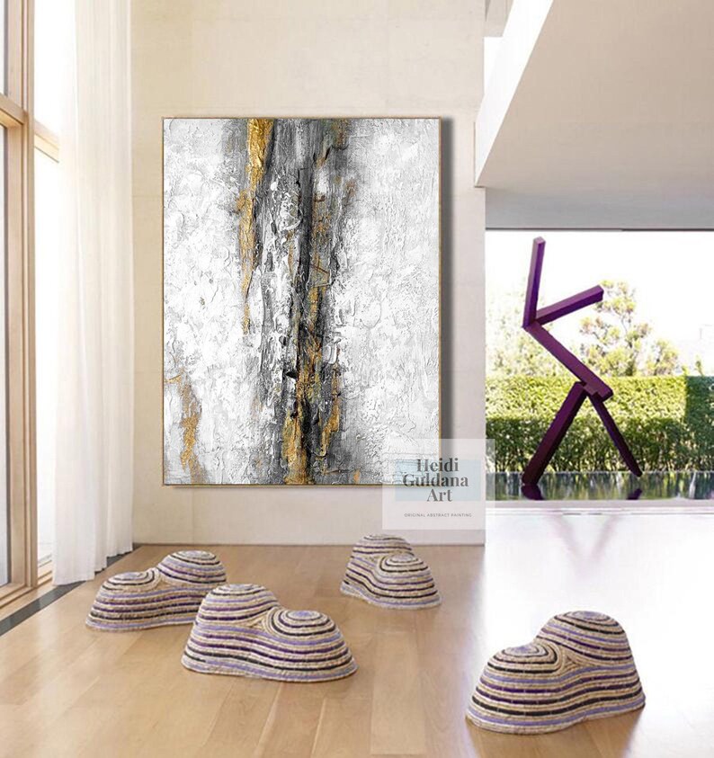 May include: Abstract painting in shades of white, gray, and gold with black accents. The painting is hanging on a white wall in a modern interior with a purple metal sculpture in the background. There are four woven, purple and white, round, decorative objects on the floor.