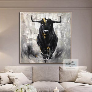 May include: Abstract painting of a black bull with gold accents against a gray and white textured background. The artwork is framed and displayed above a sofa. The artist's name, Heidi Guldana Art, is visible in the lower right corner.