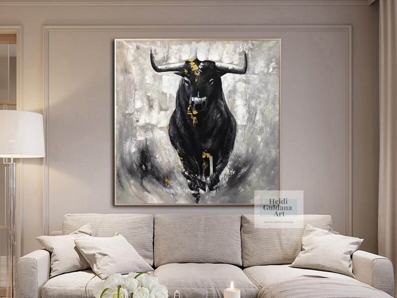 May include: Abstract painting of a black bull with gold accents against a gray and white textured background. The artwork is framed and displayed above a sofa. The artist's name, Heidi Guldana Art, is visible in the lower right corner.