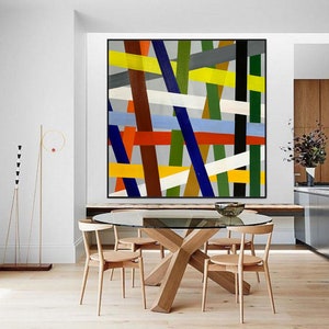 May include: Abstract geometric painting with a colorful design of intersecting lines in shades of blue, green, yellow, red, white, brown, and black. The painting is framed in a black frame and is hanging on a white wall above a wooden table.