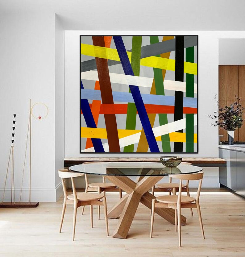 May include: Abstract geometric painting with a colorful design of intersecting lines in shades of blue, green, yellow, red, white, brown, and black. The painting is framed in a black frame and is hanging on a white wall above a wooden table.