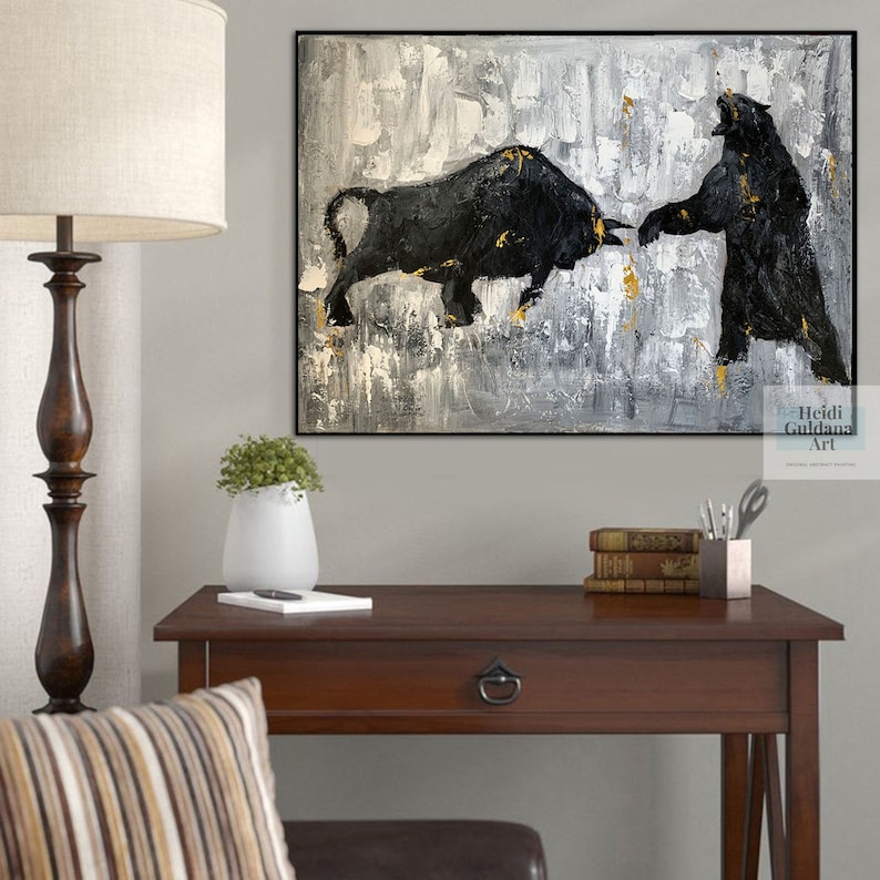 May include: Abstract painting of a black bull and bear facing each other on a gray and gold background. The painting is framed and hanging on a wall above a wooden desk.