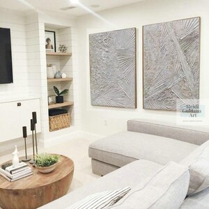 May include: Two large abstract paintings in shades of gray and white hang on a white wall above a gray sectional sofa. The paintings have a textured, layered look and are framed in a light wood.