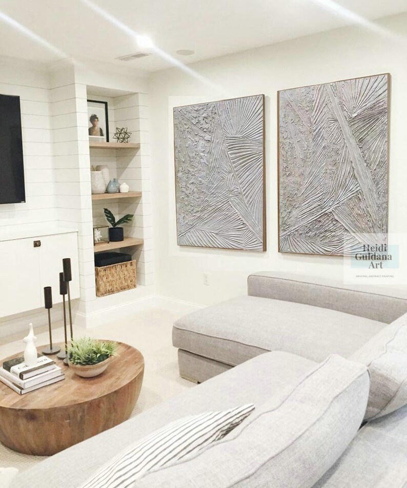 May include: Two large abstract paintings in shades of gray and white hang on a white wall above a gray sectional sofa. The paintings have a textured, layered look and are framed in a light wood.