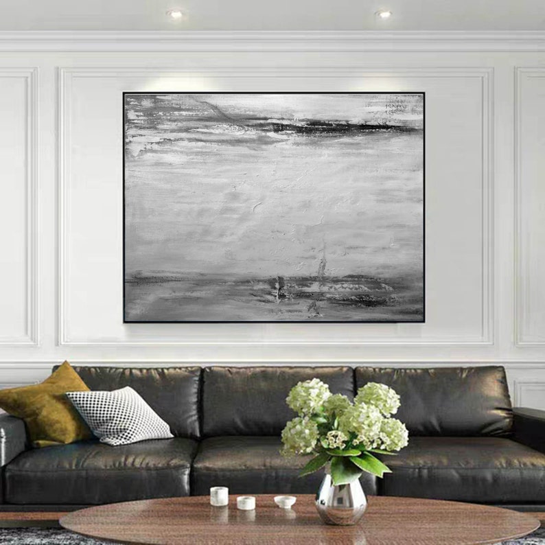 Op de afbeelding: Een zwart-wit abstract schilderij met een horizontale compositie. Het schilderij heeft een lichtgrijze achtergrond met donkergrijze en zwarte penseelstreken. Het schilderij is ingelijst in een zwart frame en hangt aan een witte muur boven een zwarte leren bank.