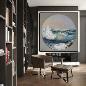 May include: A large, framed painting of a circular seascape with crashing waves and a blue sky. The painting is hanging on a wall in a modern living room with a brown leather armchair and ottoman.