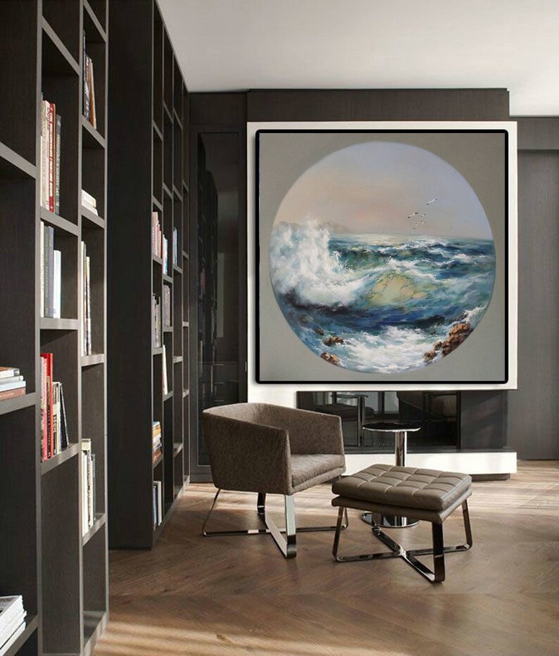 May include: A large, framed painting of a circular seascape with crashing waves and a blue sky. The painting is hanging on a wall in a modern living room with a brown leather armchair and ottoman.