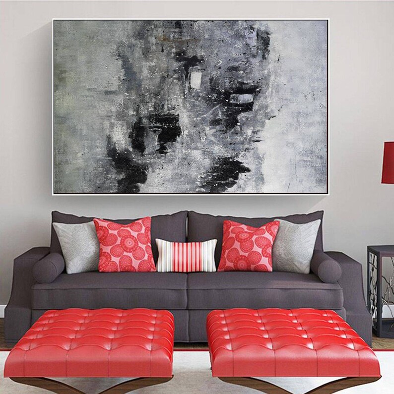 May include: A grey and black abstract painting hangs above a grey sofa with red and white patterned throw pillows. Two red tufted ottomans sit in front of the sofa.