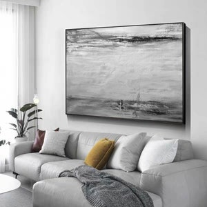 Op de afbeelding: Een zwart-wit abstract schilderij met een horizontaal landschapsontwerp. Het schilderij is ingelijst in een zwart frame en hangt aan een witte muur boven een grijze bank.