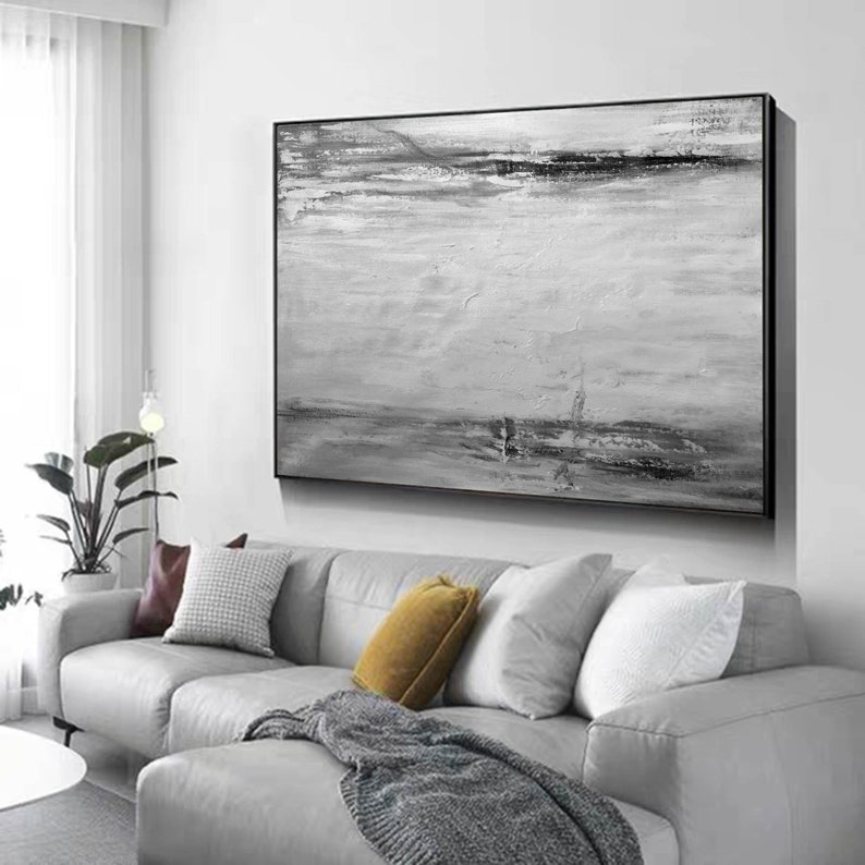 Op de afbeelding: Een zwart-wit abstract schilderij met een horizontaal landschapsontwerp. Het schilderij is ingelijst in een zwart frame en hangt aan een witte muur boven een grijze bank.
