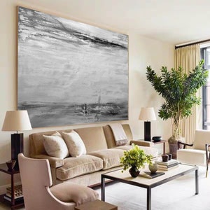Op de afbeelding: Een groot abstract schilderij in grijstinten en zwart hangt aan een muur in een woonkamer. Het schilderij heeft een textuur, gelaagde stijl met subtiele penseelstreken. Het schilderij wordt getoond boven een beige bank met kussens en een salontafel met een vaas met groen.