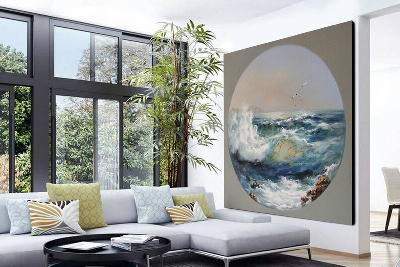 May include: A circular painting of a seascape with crashing waves and a blue sky with white clouds. The painting is framed in a light gray frame and is hanging on a wall in a living room.
