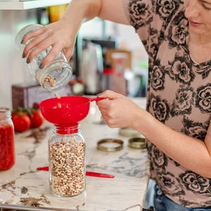 May include: A person is pouring dried beans from a glass jar into a red plastic funnel. The funnel is positioned over a clear glass jar filled with beans. The beans are a mix of black-eyed peas and brown beans.
