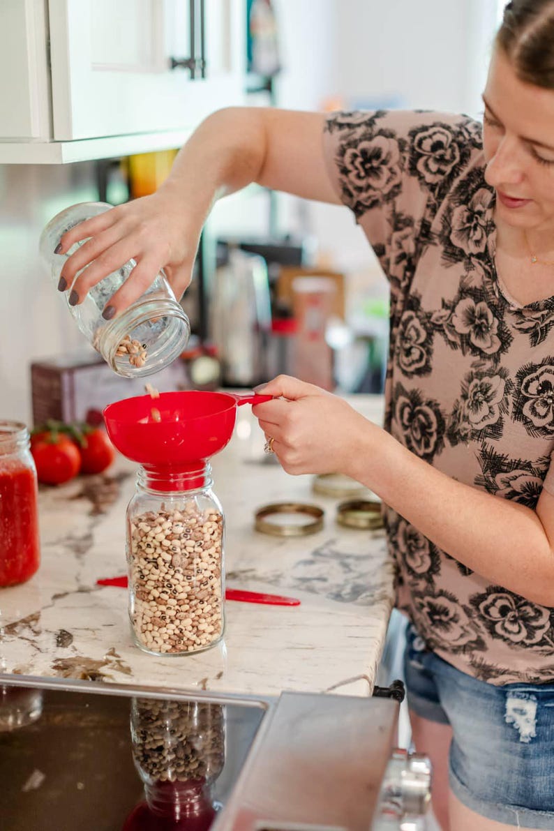 May include: A person is pouring dried beans from a glass jar into a red plastic funnel. The funnel is positioned over a clear glass jar filled with beans. The beans are a mix of black-eyed peas and brown beans.