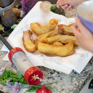 Puede incluir: Una toalla de papel blanca con varios churros de color marrón dorado espolvoreados con azúcar en polvo. Una mano está espolvoreando más azúcar en polvo desde un pequeño colador de metal.