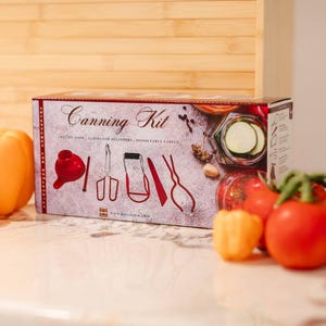 May include: A red and white canning kit box with a variety of tools for preserving food. The box has a picture of a jar of pickles and a variety of tools. The box is sitting on a counter with a variety of peppers and tomatoes.