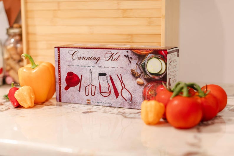 May include: A red and white canning kit box with a variety of tools for preserving food. The box has a picture of a jar of pickles and a variety of tools. The box is sitting on a counter with a variety of peppers and tomatoes.
