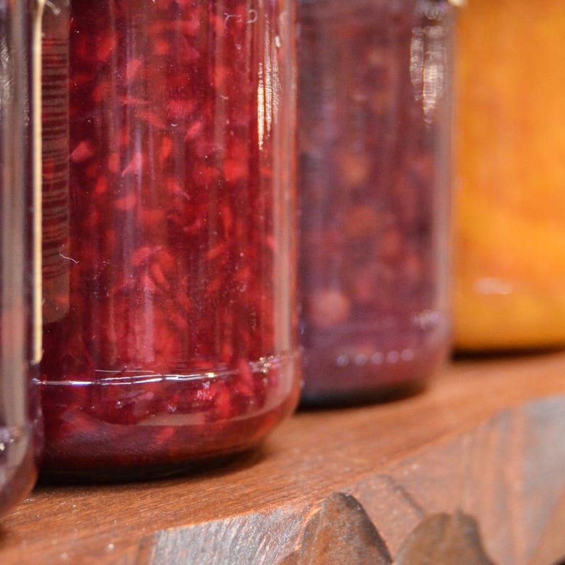 May include: Three glass jars filled with red, purple, and orange preserves. The jars are sitting on a wooden surface.