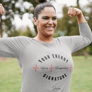 May include: A woman wearing a grey long-sleeved shirt with a black and red graphic that says "Your Energy Signature" with a red heartbeat graphic and the text "Yes Frequency".