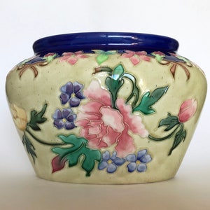 May include: A cream-colored ceramic planter with a blue rim and a floral design. The flowers are pink, yellow, and purple with green leaves.