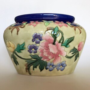 May include: A cream-colored ceramic planter with a blue rim and a floral design. The flowers are pink, yellow, blue, and green.