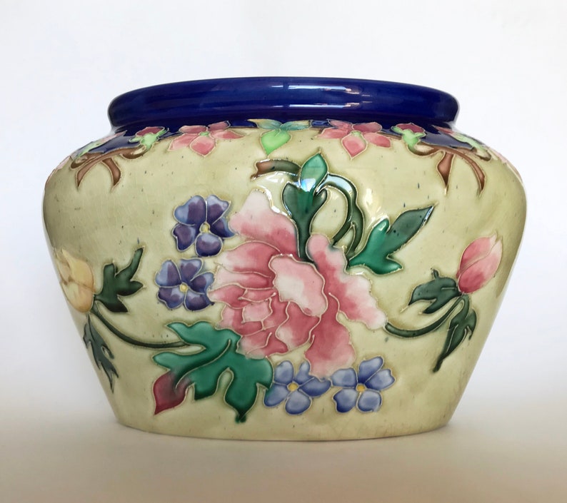 May include: A cream-colored ceramic planter with a blue rim and a floral design. The flowers are pink, yellow, and purple with green leaves.