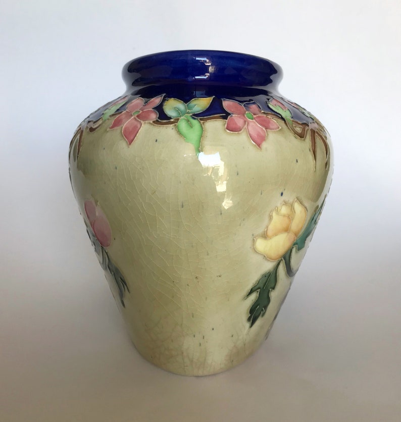 May include: A ceramic vase with a blue rim and floral designs in shades of pink, yellow, and green. The vase has a crackled glaze and a light beige background.