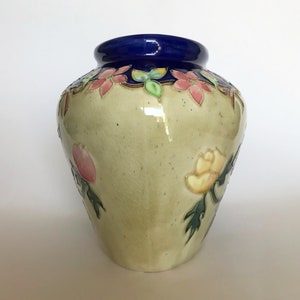 May include: A ceramic vase with a light green glaze and floral designs in shades of pink, yellow, and green. The vase has a blue rim and a slightly rounded shape.