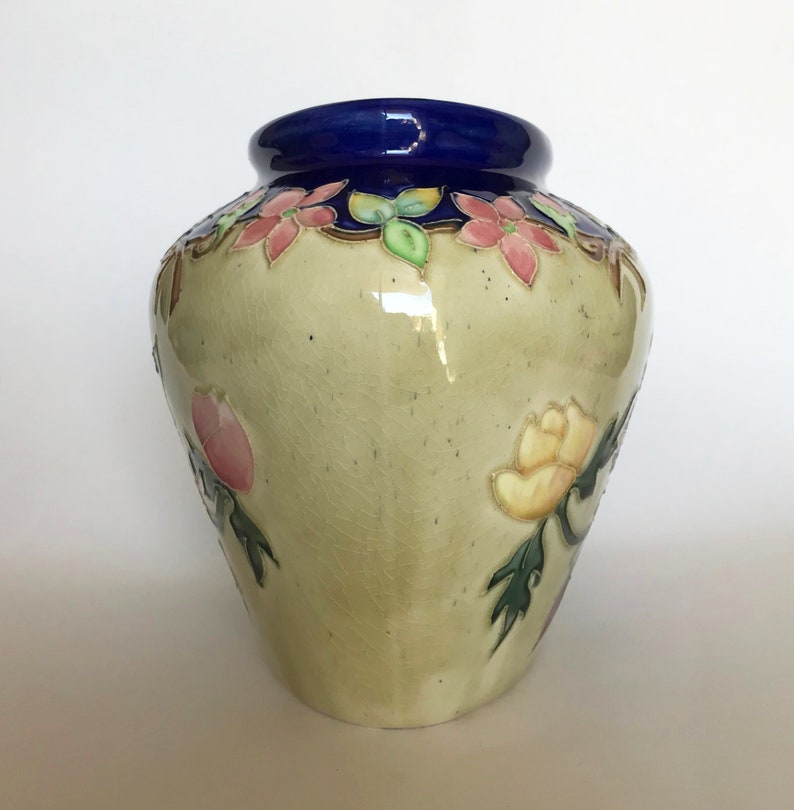 May include: A ceramic vase with a light green glaze and floral designs in shades of pink, yellow, and green. The vase has a blue rim and a slightly rounded shape.