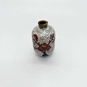 Puede incluir: Un pequeño jarrón de porcelana blanca con un diseño floral en rojo, verde y azul. El jarrón tiene un borde dorado y una forma redonda.