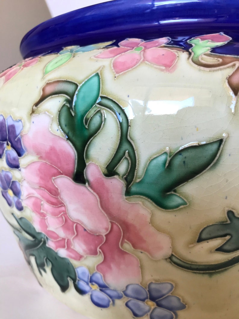 May include: A white ceramic planter with a blue rim and floral design. The planter features pink, blue, and green flowers with intricate details.
