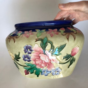 May include: A large, cream-colored ceramic planter with a blue rim. The planter is decorated with a floral design featuring pink, purple, and green flowers.