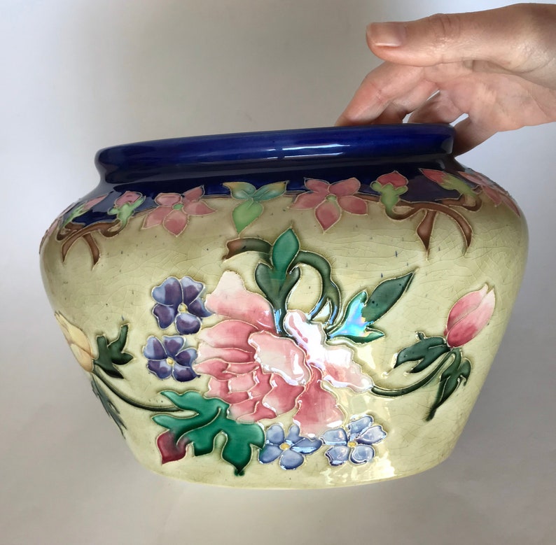 May include: A large, cream-colored ceramic planter with a blue rim. The planter is decorated with a floral design featuring pink, purple, and green flowers.