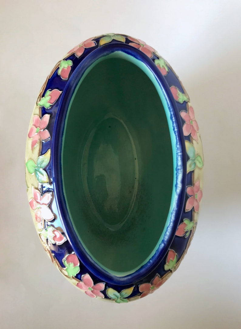 May include: An oval ceramic planter with a blue glaze and floral design. The planter has a light green interior and is decorated with pink, yellow, and green flowers.