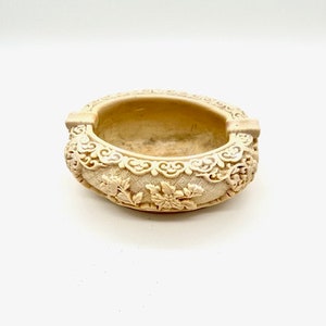 May include: A cream-colored ceramic ashtray with intricate floral and scrollwork designs.