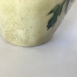 May include: A cream-colored ceramic vase with a crackled glaze and green leaf designs. The vase has a rounded bottom and a slightly flared opening.