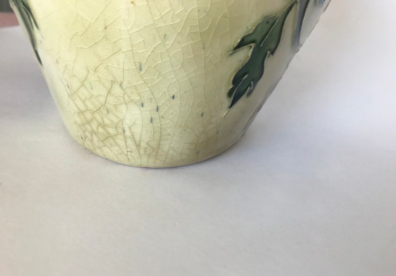 May include: A cream-colored ceramic vase with a crackled glaze and green leaf designs. The vase has a rounded bottom and a slightly flared opening.