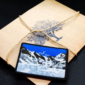 May include: A blue and white embroidered patch depicting a snowy mountain scene. The patch is surrounded by a black border and is attached to a brown paper envelope.