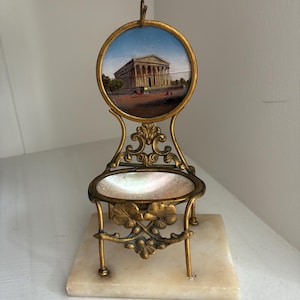 May include: An antique soap dish with a gold-coloured metal frame and a marble base. The dish features a round painting of a building with columns. The dish has an ornate design with floral accents.