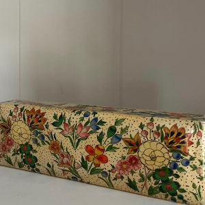 May include: A rectangular box with a cream-coloured background, decorated with a floral pattern. The flowers are in yellow, red, pink, and blue, with green leaves. The box is decorated with small black dots.