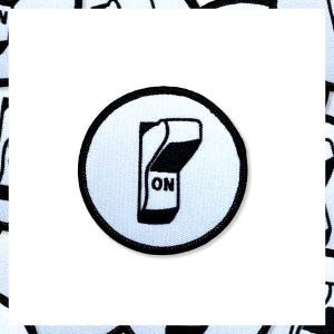 May include: A circular patch with a black border and a white background. The patch features a black and white illustration of a switch in the "ON" position. The word "ON" is printed in black.