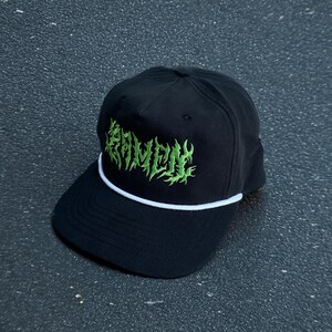 May include: A black baseball cap featuring the word RAMEN embroidered in a bright green, stylized, jagged font across the front panel. The cap includes a white cord detail running horizontally across the base of the crown above the brim.