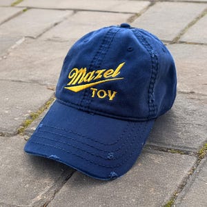 May include: A navy blue baseball cap with the word "Mazel" in yellow script and "TOY" in yellow block letters. The cap has a distressed look with frayed edges.