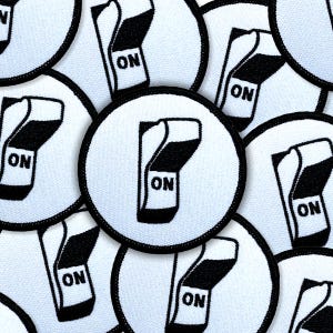 May include: White circular patches with a black border and a black and white graphic of a switch in the "ON" position. The word "ON" is printed in black on the switch. The patches are made of a textured fabric.