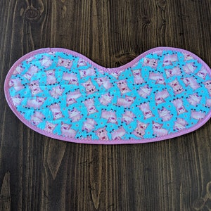 May include: A pink and blue fabric bib with a pattern of pink pigs on a light blue background. The bib has a pink trim.